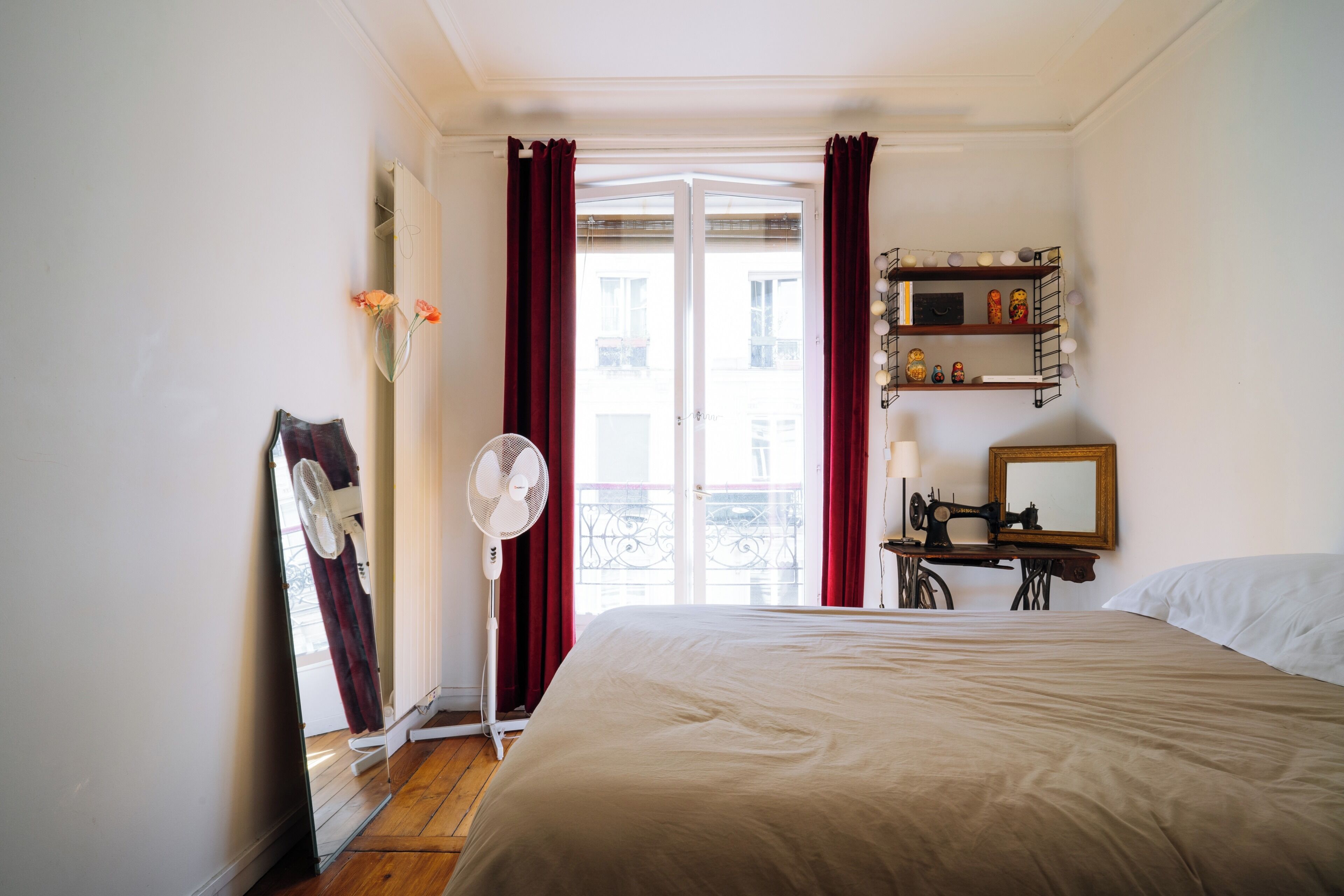 A hotel room with a bed, a fan, a mirror, a sewing machine, and a window with curtains.