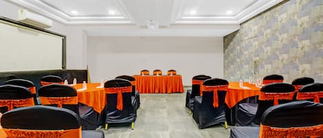 Un salón de conferencias con filas de mesas, sillas negras con cintas naranjas y una pantalla de proyector en la pared izquierda.