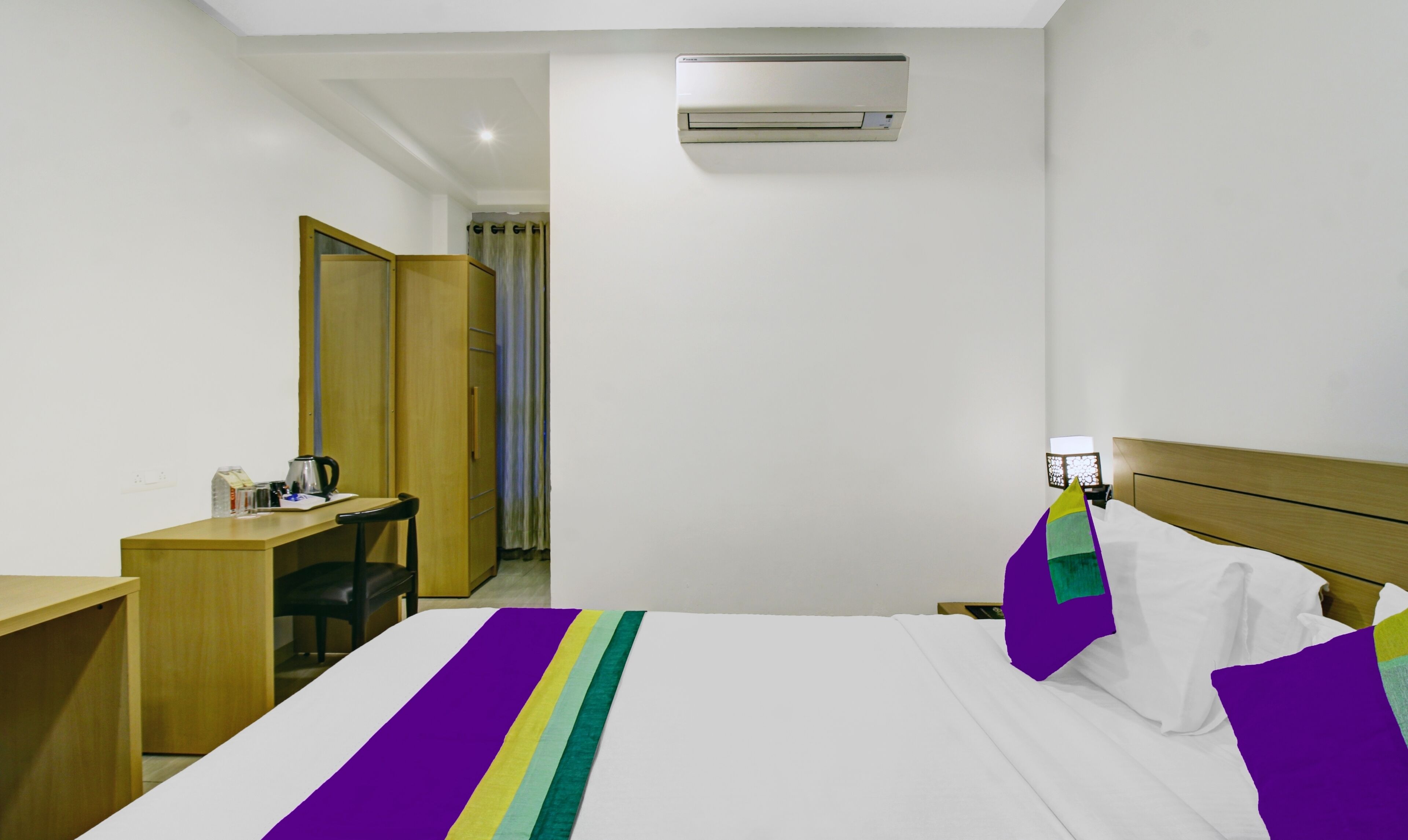 A hotel room with a bed, a desk, a chair, an air conditioner, and a nightstand with a lamp.