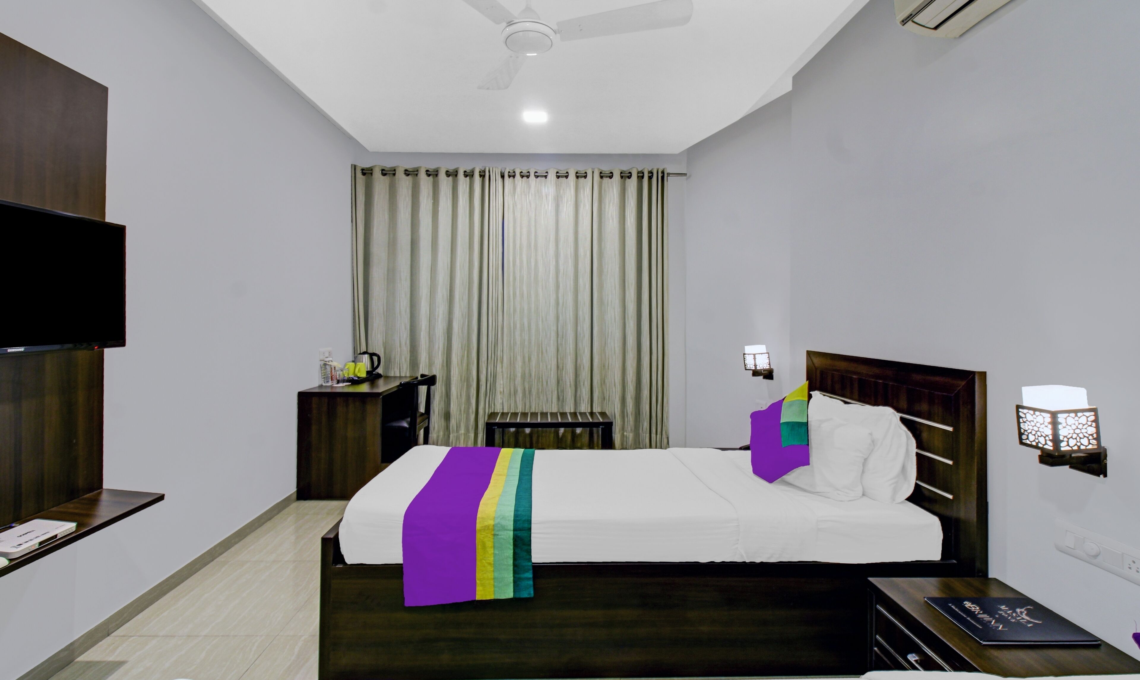 A hotel room with a bed, a TV, a desk, a chair, a nightstand, a lamp, and a ceiling fan.