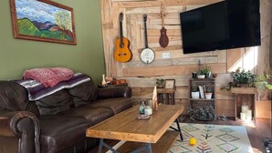 Uma sala de estar aconchegante com um sofá de couro marrom, uma mesa de centro de madeira, uma TV fixada em uma parede de madeira e diversos instrumentos musicais e objetos decorativos.