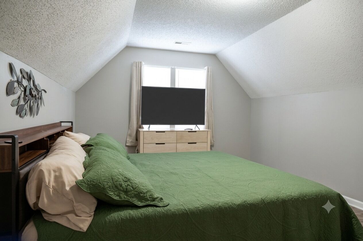 A bedroom with a sloped ceiling, a bed with green bedding, a bedside table, a dresser, and a window with curtains.