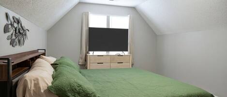 A bedroom with a sloped ceiling, a bed with green bedding, a bedside table, a dresser, and a window with curtains.