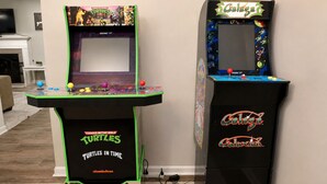 Dos máquinas de videojuegos arcade, una con Teenage Mutant Ninja Turtles y otra con Galaxian, en una habitación con chimenea y televisor.