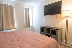 A bedroom with a bed, a television mounted on the wall, a desk with a lamp, and a window with curtains.