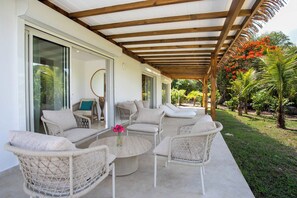 A covered outdoor seating area with wicker furniture, a glass door entrance, and a view of a garden with tropical plants. - Encore (8 bedrooms) - Lovely villa with pool, Terres Basses, St. Martin (Terres Basses)