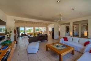 A spacious living area with a sofa, ottoman, and coffee table. There is a kitchen area in the background, and a dining area with chairs.