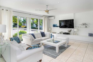 A modern living room with white furniture, a flat-screen TV, and a view of greenery outside. - Encore (8 bedrooms) - Lovely villa with pool, Terres Basses, St. Martin (Terres Basses)