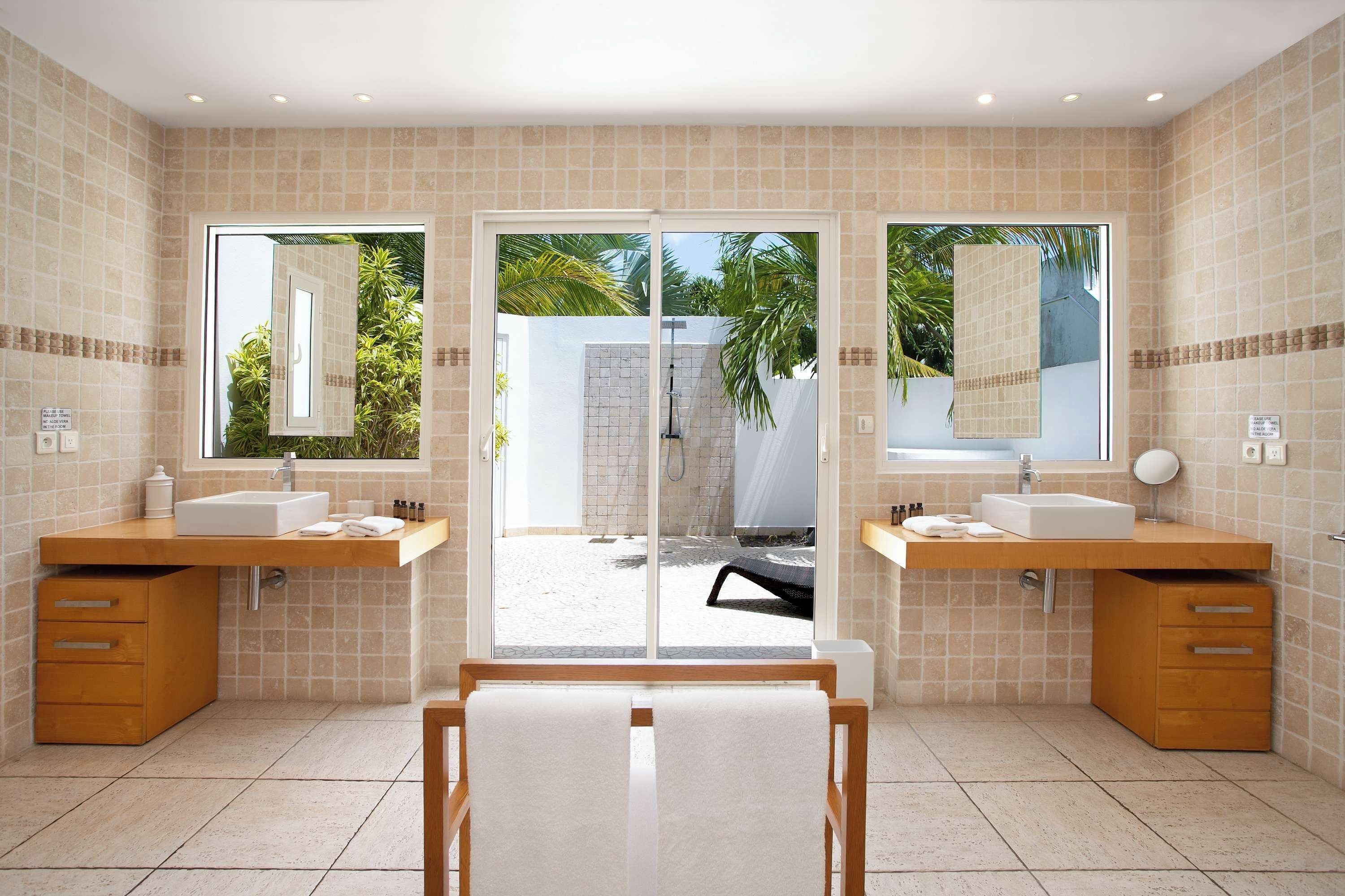 Un baño moderno con dos lavamanos, un espejo grande y vistas a una piscina y palmeras.