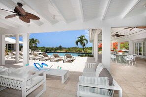 Un espacio al aire libre techado con mobiliario blanco, piscina y palmeras.