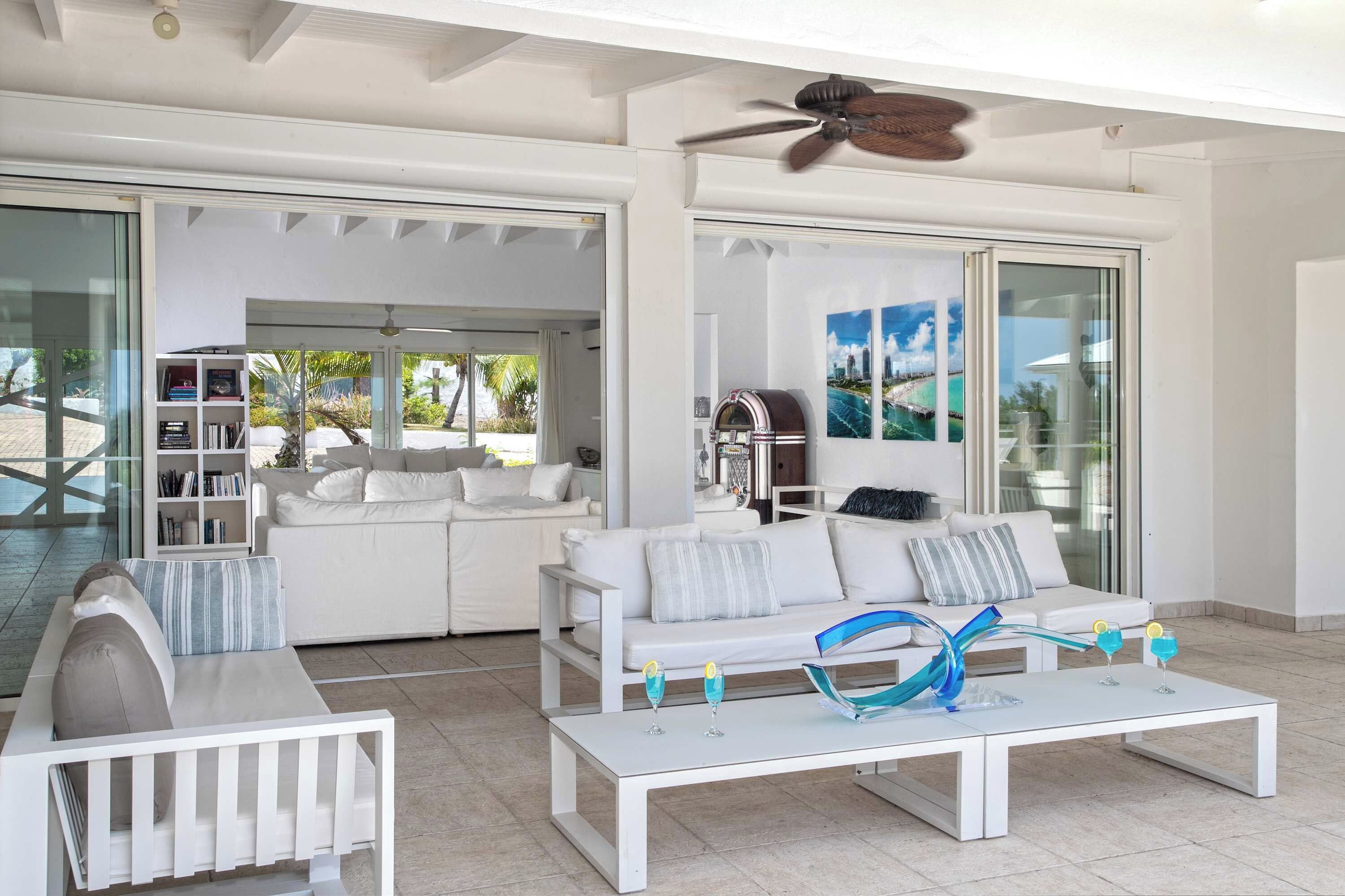 Un moderno salón con muebles blancos, un ventilador de techo y un ventanal que da a una zona de piscina.