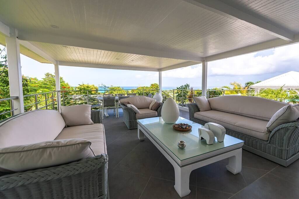 A covered outdoor seating area with wicker furniture and a glass coffee table, overlooking a scenic view.