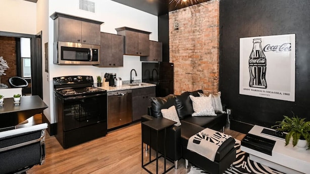A modern kitchen with dark cabinetry, a brick accent wall, and a large poster of Andy Warhol's Coca-Cola artwork.