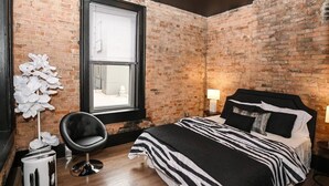 A bedroom with a brick wall, a large window, a black chair, a bed with striped bedding, and a bedside table with a lamp.