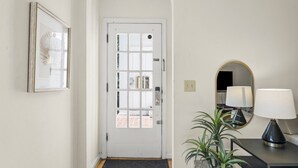 A hallway with a white door, a framed picture, a potted plant, a mirror, and a lamp on a console table. - Zan Court Unit 1 · Mt Lookout Hideaway (Cincinnati)