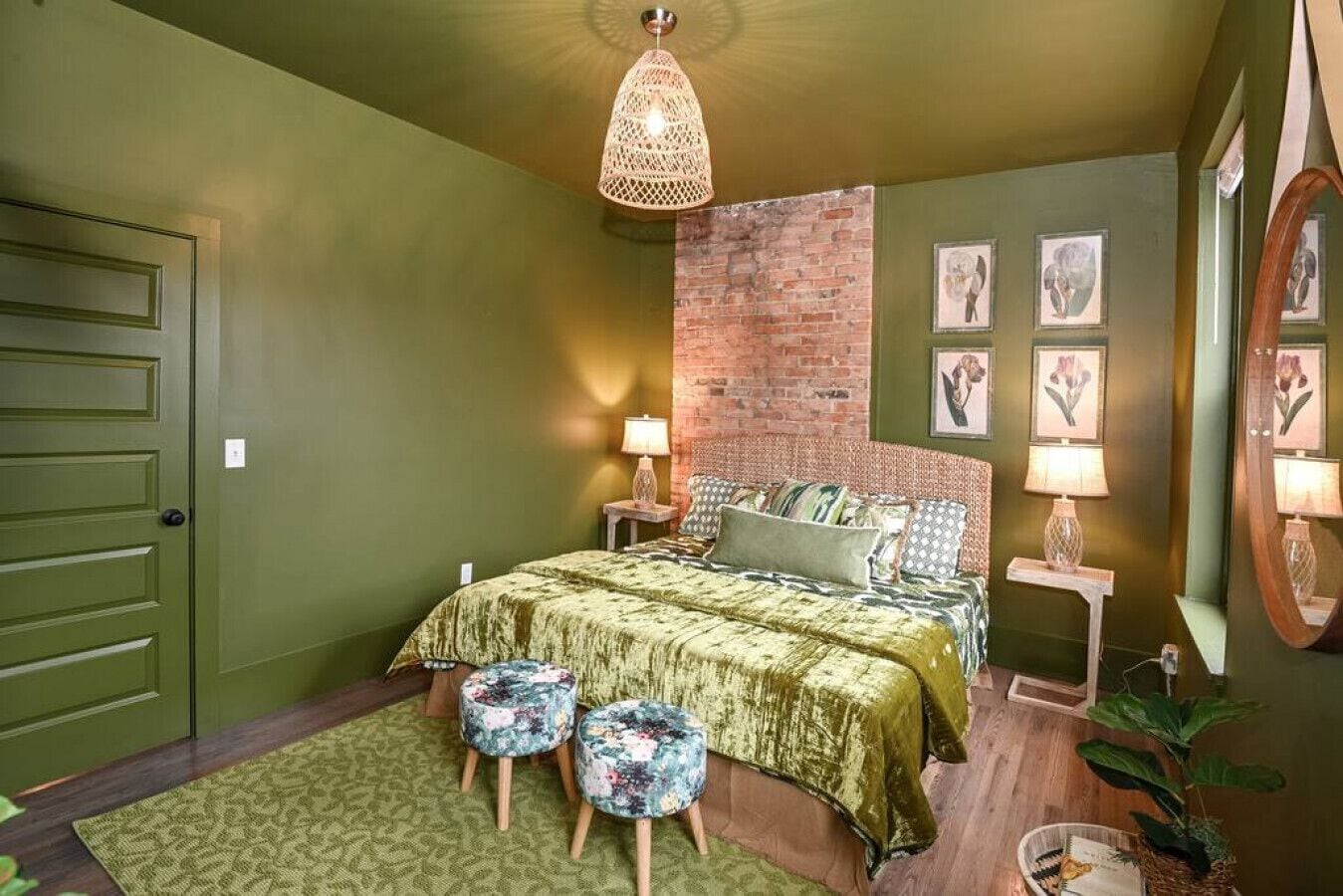 A bedroom with a green color scheme, a brick accent wall, a bed with floral-patterned bedding, two bedside lamps, and a hanging light fixture.