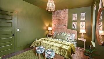 A bedroom with a green color scheme, a brick accent wall, a bed with floral-patterned bedding, two bedside lamps, and a hanging light fixture.