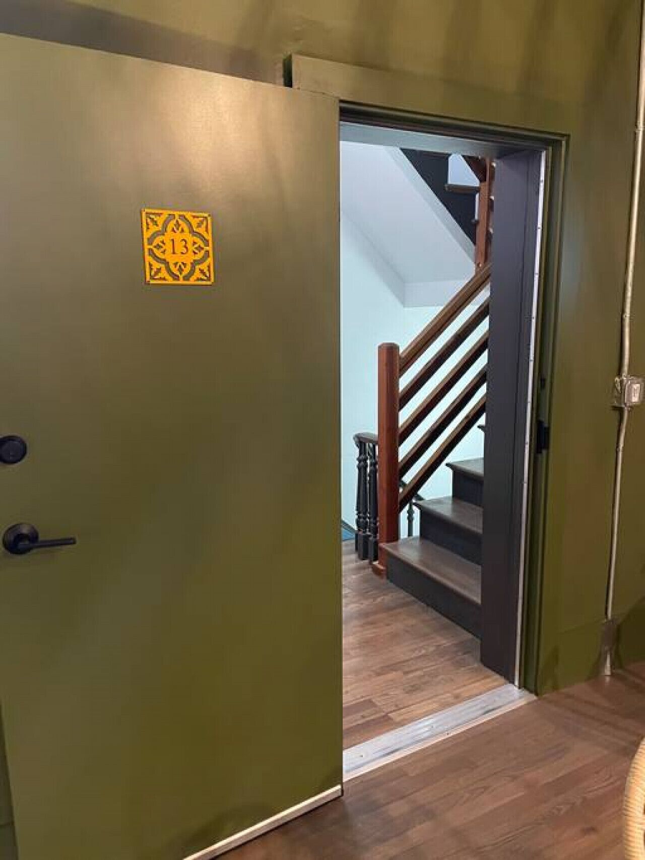 Una porta verde con un cartello giallo, una scala di legno e una parete di colore chiaro.