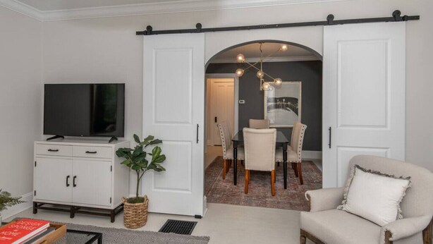 A modern living room with a TV, white cabinets, a plant, and a dining area with a chandelier. - Lower Cambridge · Cozy Cambridge (Cincinnati)