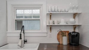 A kitchen with a white sink, a window with blinds, a coffee maker, and shelves with glassware and dishes. - Lower Cambridge · Cozy Cambridge (Cincinnati)