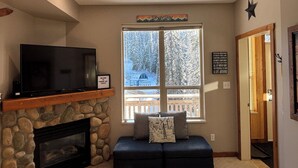 A cozy living room with a stone fireplace, a flat-screen TV on a wooden shelf, a bench with cushions, and a large window with a view of a snowy landscape. - Perfect Ground Floor Condo in Creekside - ski right from your door! (Silver Star Mountain)
