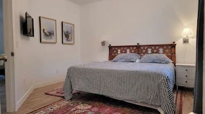 Um quarto com cama, mesa de cabeceira, luminária e televisão na parede.