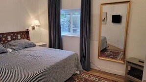 Un dormitorio con cama, mesita de noche, lámpara, ventana con cortinas, espejo y una pequeña mesa.