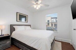 A bedroom with a bed, a nightstand, a ceiling fan, a window with a view, and a door.