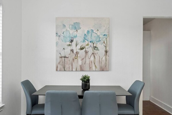 A dining area with a table and chairs, a wall-mounted painting, and a small potted plant.