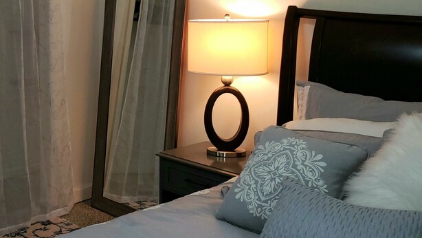A neatly made bed with a pillow, a bedside table with a lamp, and a mirror on the wall.