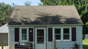 A small, single-story house with a dark roof, white siding, and red shutters. It has a front door with a number '3040' above it, a small porch with a bench, and a wooden fence in front.