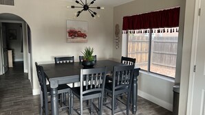 A dining area with a dark wooden table, six chairs, a black chandelier, a red truck painting, and a window with a red valance. - New family retreat w/private heated pool and hot tub (Chandler)