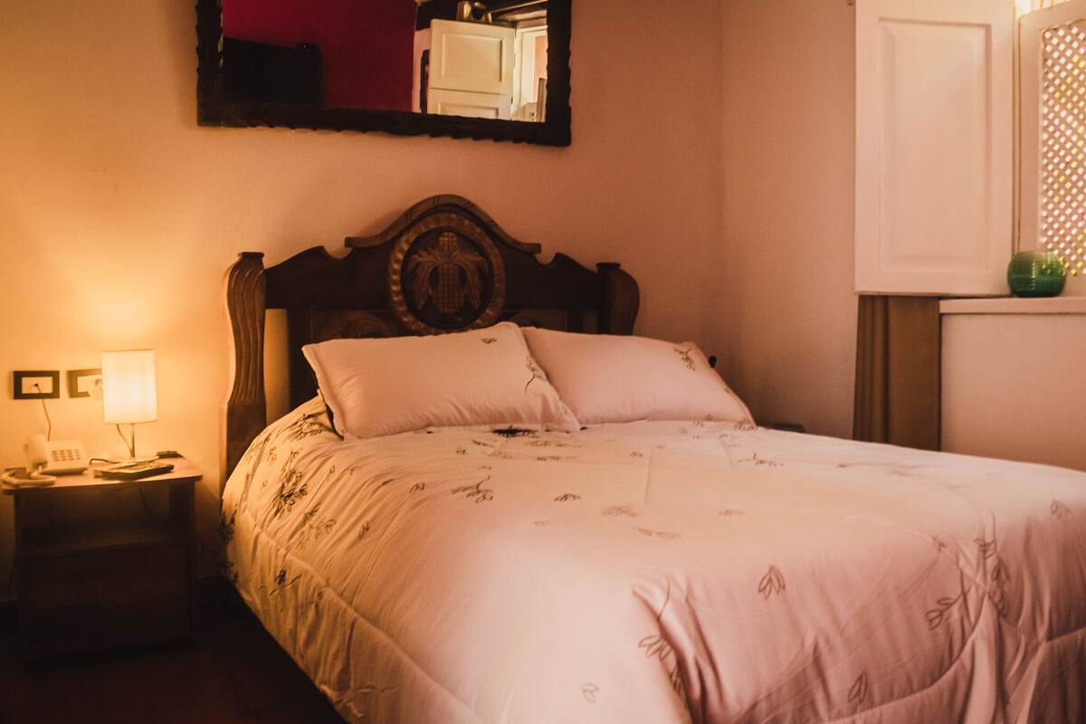 A neatly made bed with a floral bedspread and two pillows, a bedside lamp, and a mirror on the wall.
