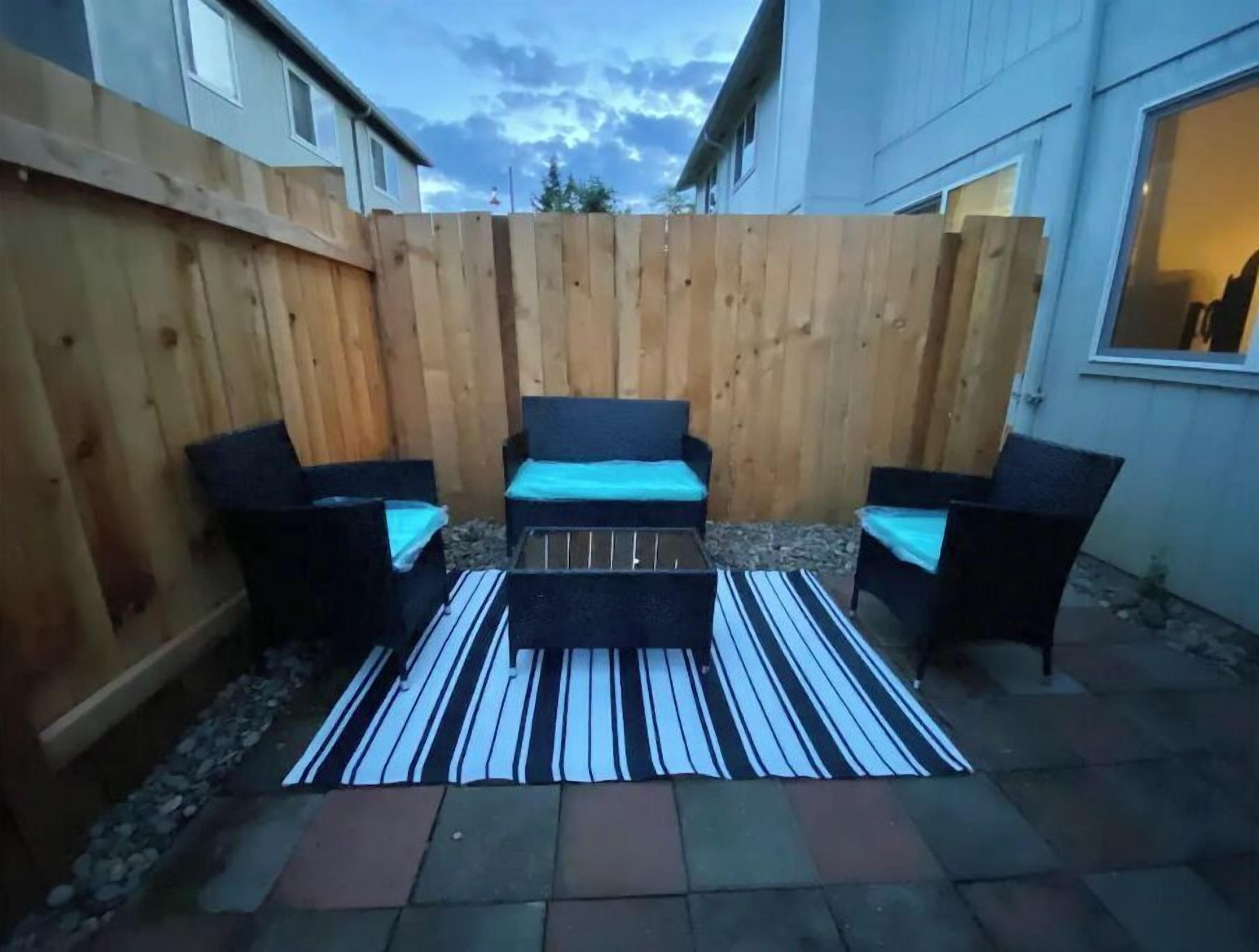 Un patio con valla de madera, dos sillas negras, un banco y una alfombra a rayas.