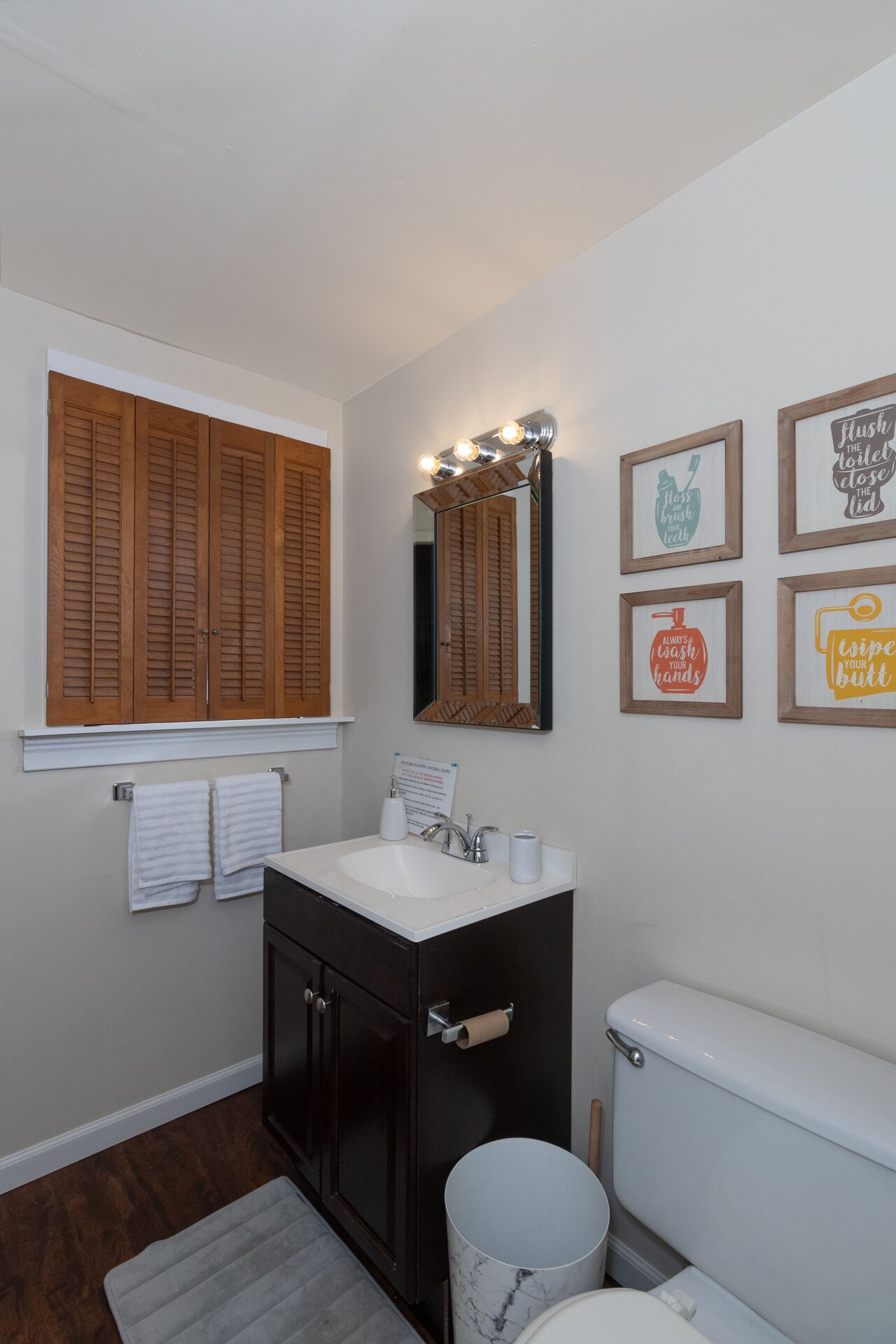 A bathroom with a vanity, a toilet, a window with wooden shutters, and wall-mounted light fixtures.