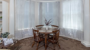 Uma área de refeições no canto, com uma mesa redonda e quatro cadeiras de madeira, cercada por cortinas brancas e um parapeito de janela em arco.
