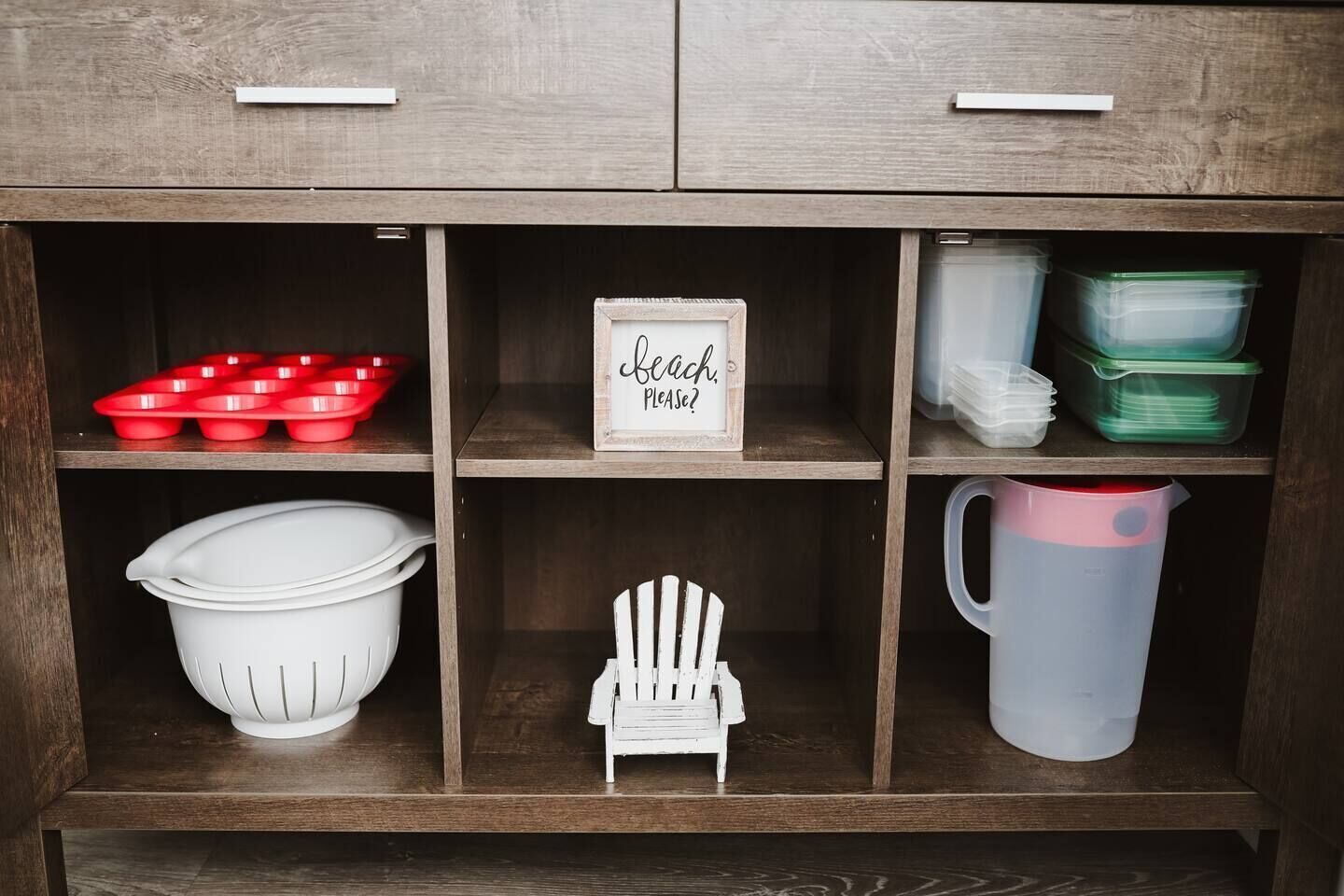 A wooden cabinet with multiple compartments, including red muffin tins, white plastic containers, a white plastic jug, and a small white chair.