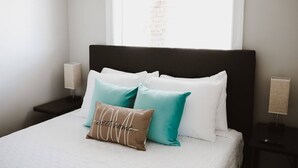 Un letto rifatto con cuscini bianchi e color acquamarina, una testata scura e abat-jour su un comodino.
