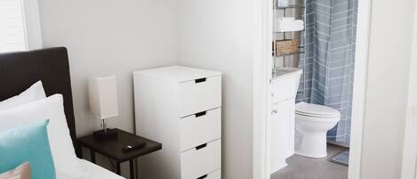Un dormitorio con cama, mesita de noche, lámpara y un mueble blanco para guardar cosas.