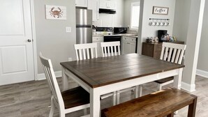 Zona pranzo con tavolo in legno e sedie bianche, cucina con mobili bianchi ed elettrodomestici in acciaio inossidabile, e angolo caffè con tazze.