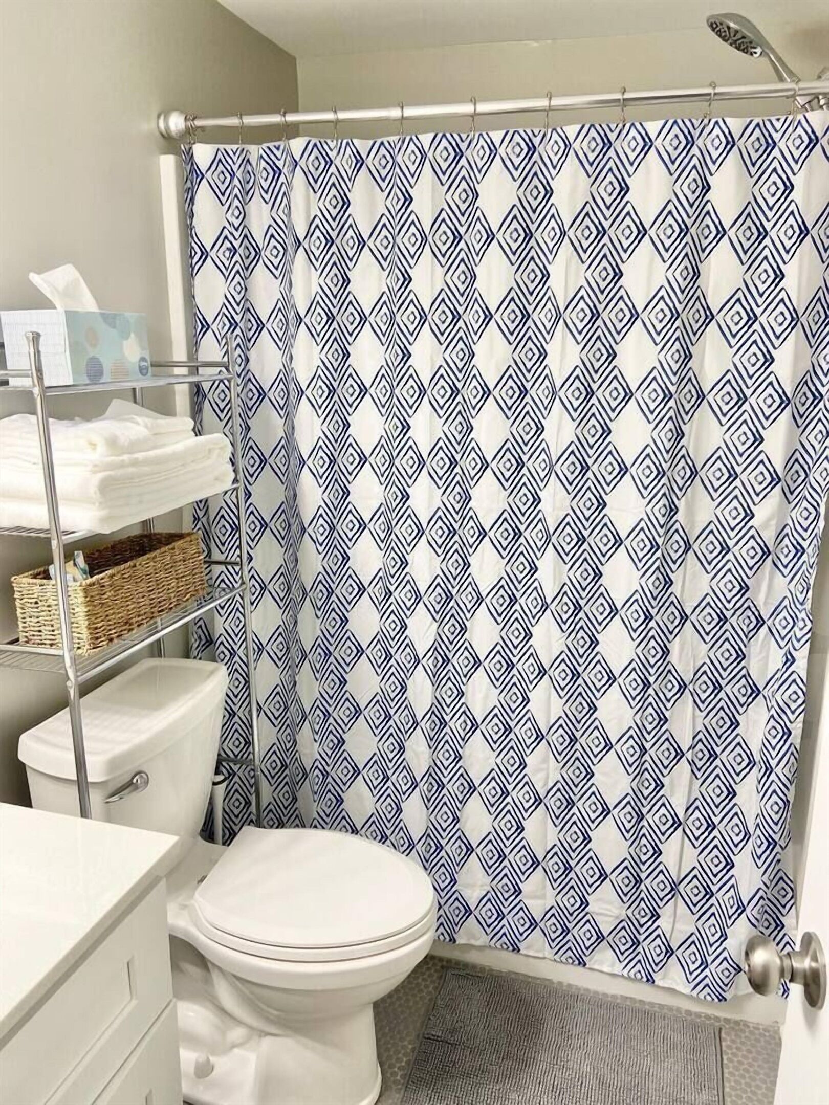 Un bagno con un water bianco, una tenda da doccia con motivo geometrico, uno scaffale in metallo con asciugamani piegati e un cesto in vimini.