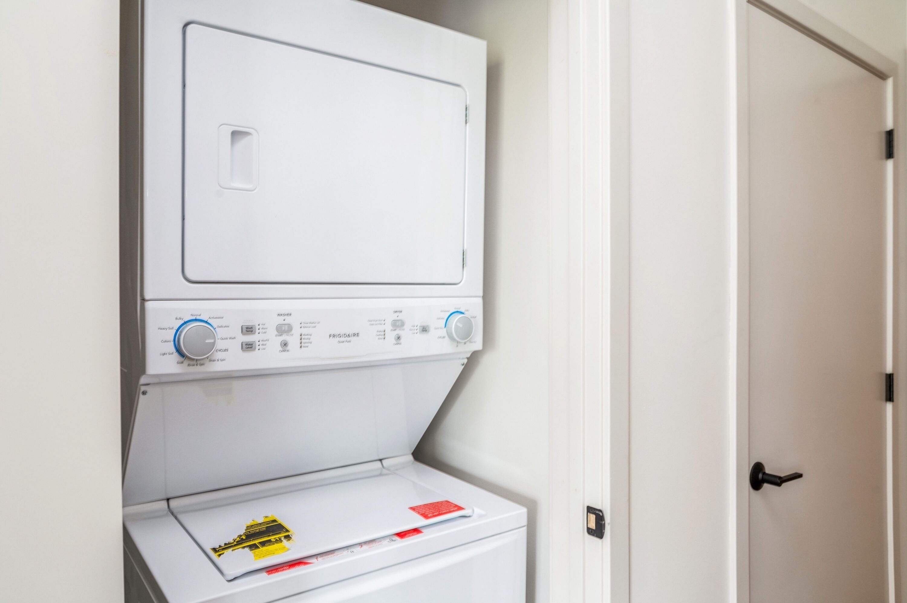 A front-loading washing machine with a control panel, a door, and a laundry basket with a yellow caution label.