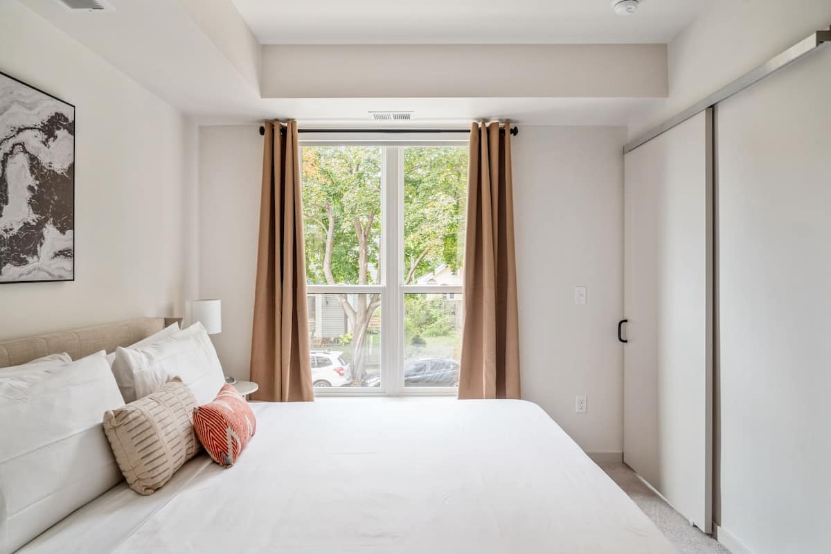Un dormitorio con una cama grande, una ventana con cortinas, una lámpara de noche y una puerta.