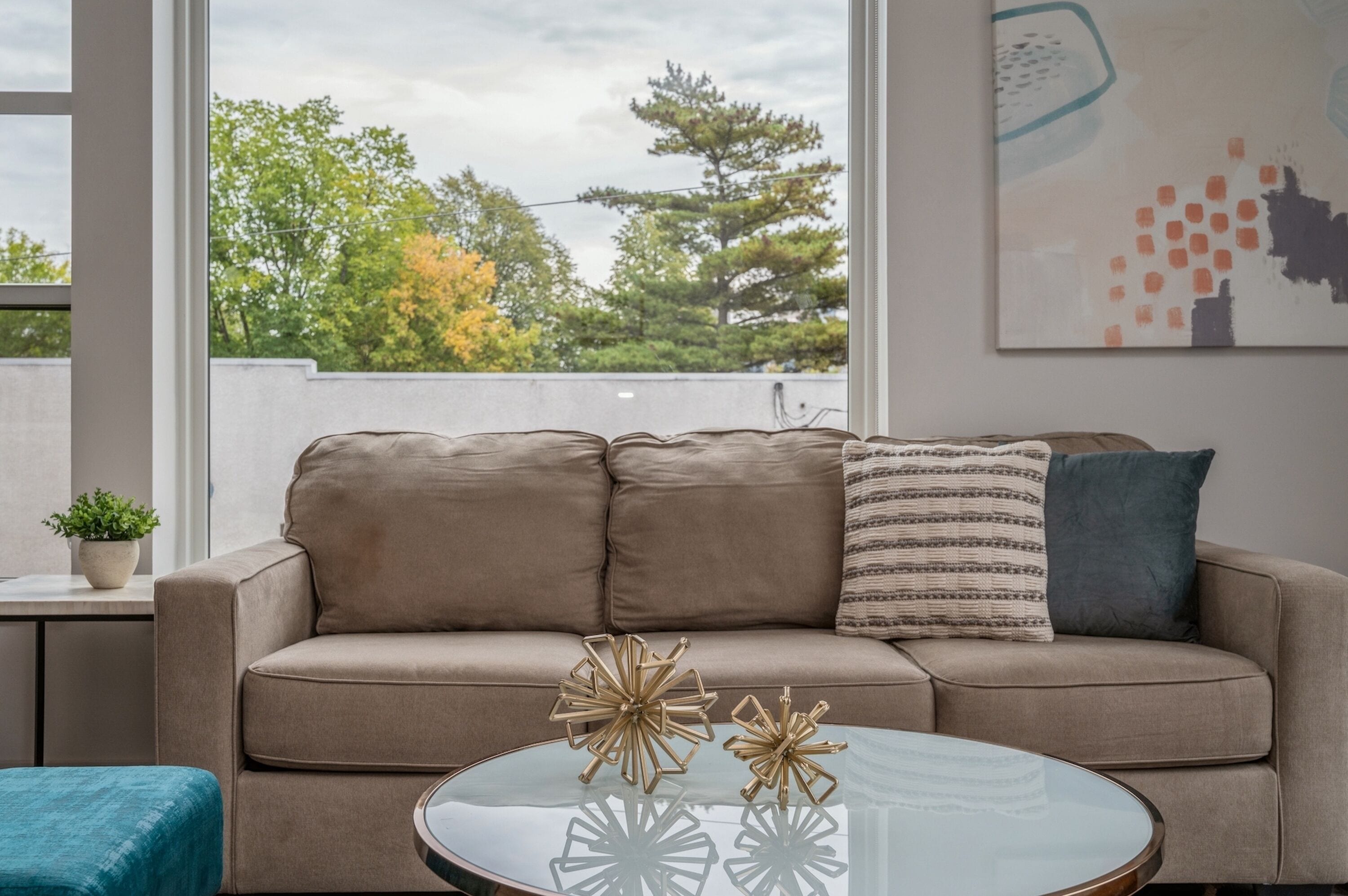 A modern living room with a beige sofa, a glass coffee table with decorative items, and a view of trees through large windows.