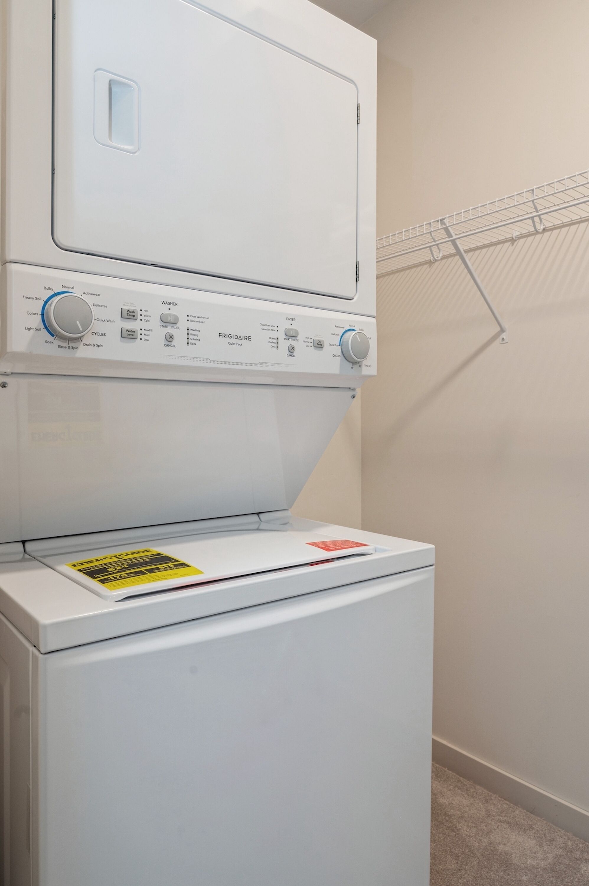 A front-loading washer and dryer unit with a control panel, a yellow caution label, and a metal drying rack mounted on the wall.