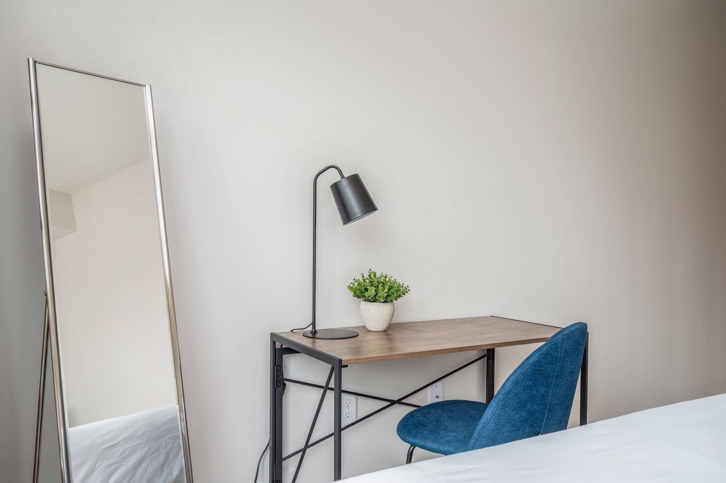 A minimalist room with a bed, a desk with a lamp, a chair, and a potted plant.