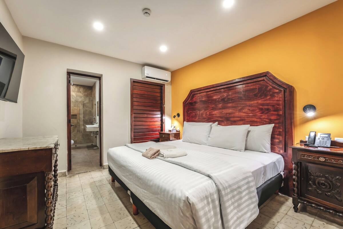 A hotel room with a large bed, wooden headboard, bedside table with a lamp, and an air conditioning unit.
