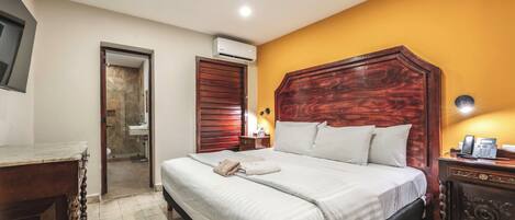Habitación de hotel con una cama grande, cabecera de madera, mesita de noche con lámpara y unidad de aire acondicionado.