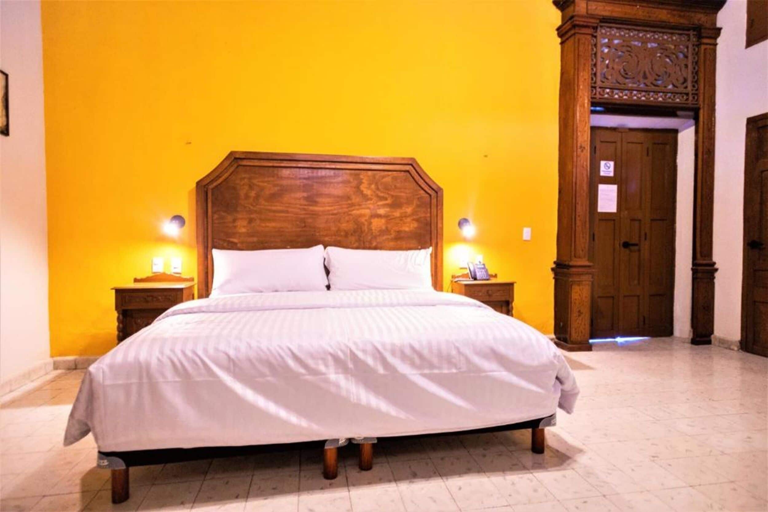 A bed in a room with a yellow wall, wooden headboard, and two bedside tables with lamps.