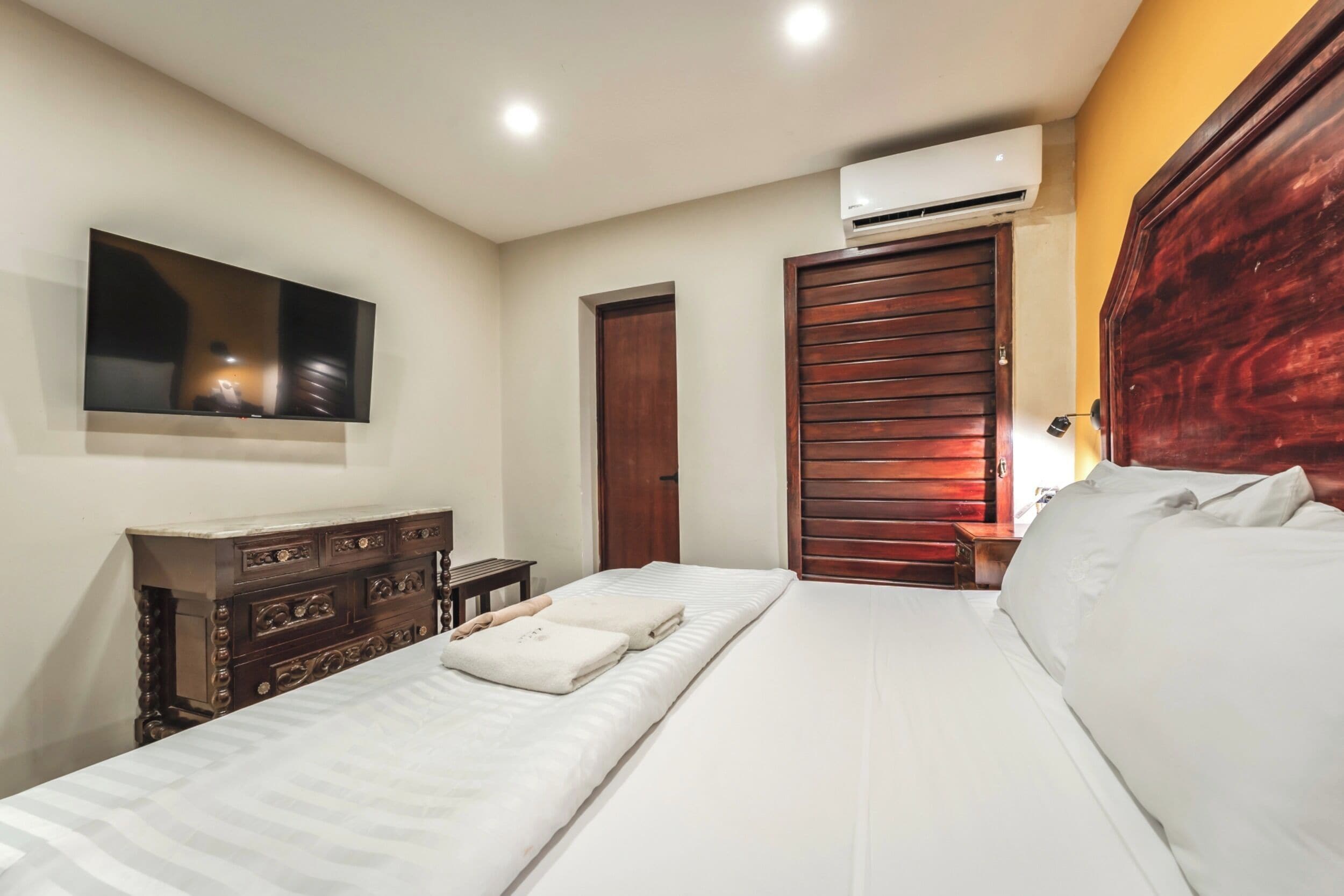 A bedroom with a bed, a dresser, a TV mounted on the wall, a wooden wardrobe, and an air conditioning unit.